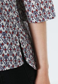 Chemise à manches courtes avec un fond crème orné de motifs losanges bleus et carrés rouges, affichant une coupe légèrement ample et des fentes latérales.