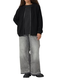 Vrouw in een zwarte ritsjas, donkere trui, wijde grijze jeans met vervaging en zwarte chunky schoenen, staand met één hand achter haar rug.