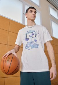 Camiseta de color gris claro con un gráfico de un aro de baloncesto, texto "SOLO HAZLO" y diseños de baloncestos coloridos. El modelo sostiene un baloncesto.