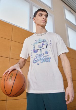 Camiseta de color gris claro con un gráfico de un aro de baloncesto, texto "SOLO HAZLO" y diseños de baloncestos coloridos. El modelo sostiene un baloncesto.