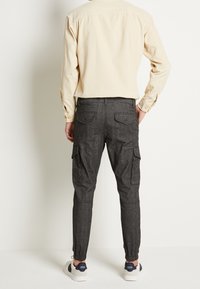 Lichtbeige herenoverhemd met knopen en lange mouwen, gecombineerd met donkergrijze cargobroeken met elastische boorden en meerdere zakken. Witte sneakers.