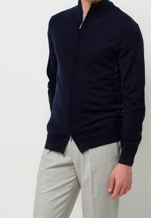 Homme portant un pull zippé bleu marine et un pantalon gris clair sur un fond uni.