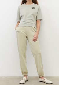 Felpa grigia chiara corta con collo rotondo e maniche corte, abbinata a pantaloni jogger verde chiaro con polsini elasticizzati e dettagli testuali.