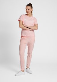 Ljust rosa bomullströja med en vit logotyp, kombinerad med matchande rosa joggingbyxor. Båda har en slät textur och en figurnära design.