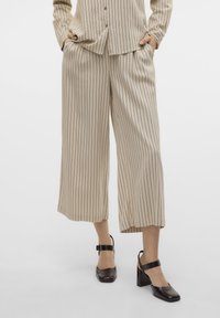 Donna che indossa pantaloni beige a gamba larga e taglio cropped con righe verticali nere e scarpe Mary Jane con tacco a blocco nero, con le mani nelle tasche.