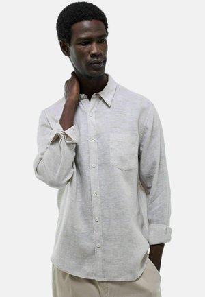 Homme à la peau foncée portant une chemise beige clair à boutons avec les manches retroussées, debout avec une main touchant son cou.