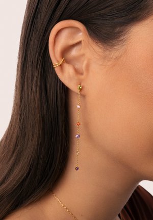 Boucles d'oreilles en or avec des pierres précieuses colorées - vertes, roses, oranges, violettes - suspendues à une chaîne délicate, sur un fond neutre.