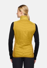 Gelbe isolierte Weste mit gestepptem Muster, hohem Kragen und seitlichen Einsätzen. Wird über einem schwarzen Langarmshirt getragen und zeigt eine figurbetonte Passform.