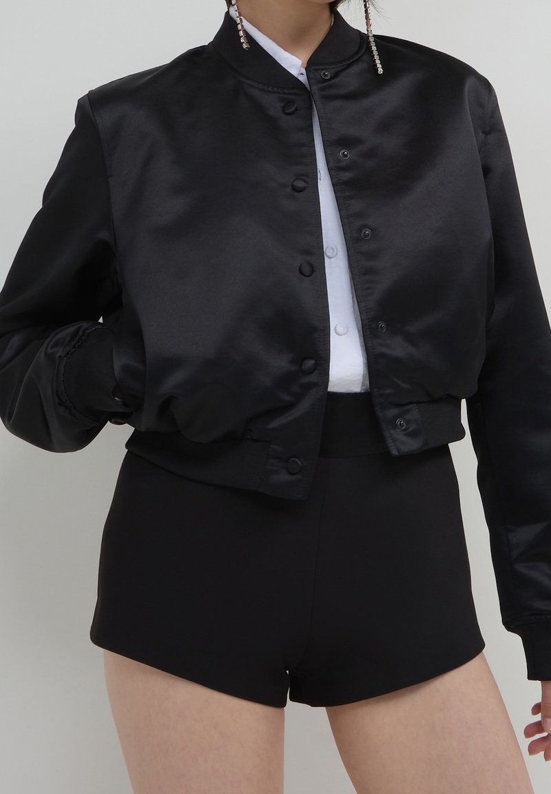 Czarna satynowa kurtka bomberka o dopasowanym kroju, z zatrzaskami i ściągaczami na rękawach, zestawiona z czarnymi szortami z wysokim stanem.