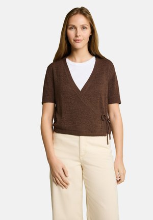 Frau trägt einen braunen kurzärmligen Wickelpullover über einem weißen Hemd und cremefarbenen Hosen, steht vor einem schlichten Hintergrund.