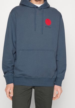 Mann trägt einen blauen Kapuzenpullover mit einem roten Kreis und japanischem Text auf der linken Brust, kombiniert mit einer dunklen Hose.