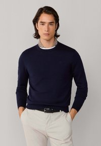 Marineblauwe gebreide trui met een ronde halslijn en geribbelde manchetten, gecombineerd met een patroonblouse met kraag eronder. De achtergrond is neutraal.