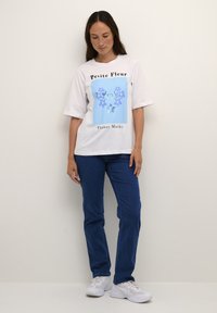 Balta marškinėliai su mėlynu gėlių grafiniu dizainu ir užrašu "Petite Fleur", derinami su mėlynomis džinsinėmis kelnių ir baltais sportiniais bateliais. Modelis stovi prieš neutralų foną.