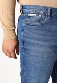 Vaqueros de denim azul con un corte recto, que presentan una pequeña etiqueta de Calvin Klein en el bolsillo trasero y una textura ligeramente deslavada.