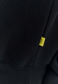 Černá mikina z materiálu s žebrovanou strukturou. Obsahuje malou žlutou nášivku se slunečním motivem všitou do bočního švu.