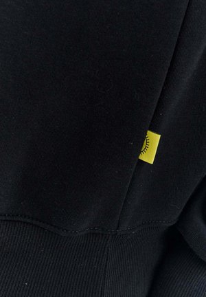 Czarna bluza z tkaniny o prążkowanej fakturze. Posiada małą żółtą metkę z wizerunkiem słońca wszytą w boczny szew.