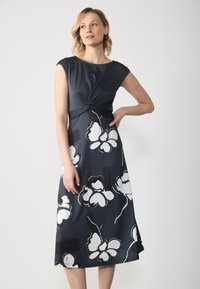 Marineblaues Kleid mit angeschnittenen Ärmeln, verdrehtem Vorderteil, floralem Druck in Weiß und Schwarz, knielang. Glatte Stoffqualität mit leichtem Glanz.