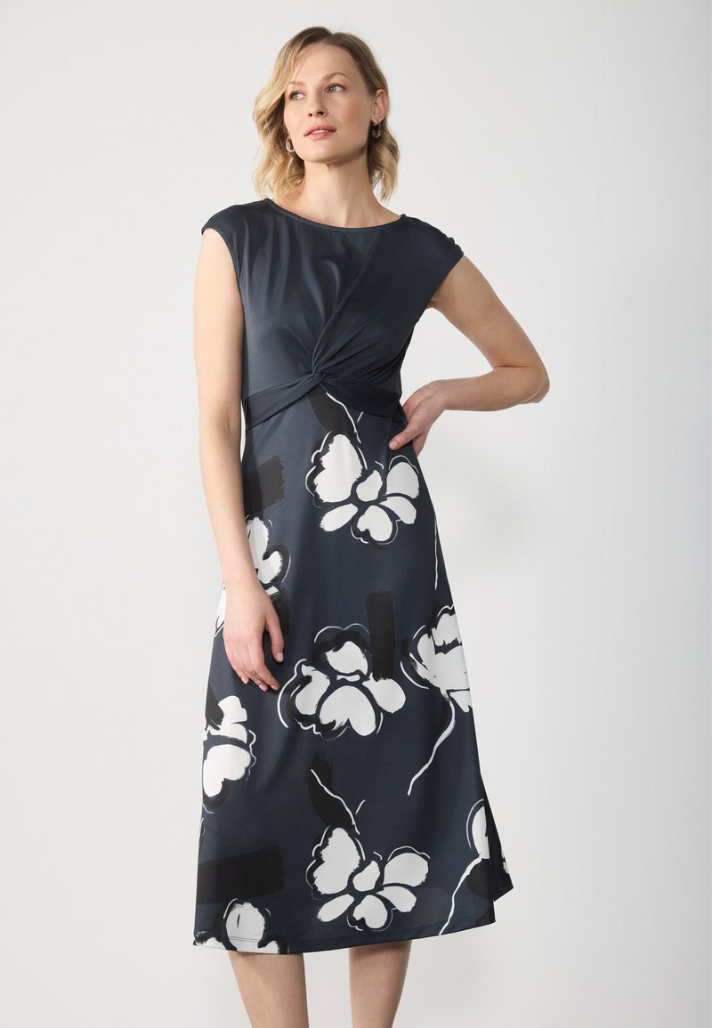 Marineblaues Kleid mit angeschnittenen Ärmeln, verdrehtem Vorderteil, floralem Druck in Weiß und Schwarz, knielang. Glatte Stoffqualität mit leichtem Glanz.