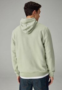 Sudadera verde claro con capucha ajustable, puños y dobladillo acanalados. Textura de tejido suave; llevada sobre una camiseta blanca en capas y jeans oscuros.