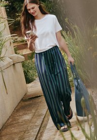Jeune femme marchant en extérieur le long d'un chemin de jardin, regardant son smartphone, portant un t-shirt blanc, un pantalon large à rayures, des sandales, et portant un sac fourre-tout bleu.