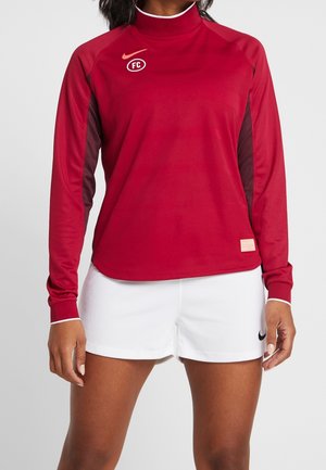 Frau trägt ein rotes langärmliges Sportoberteil mit FC-Logo und weiße Sportshorts mit einem schwarzen Nike-Swoosh.
