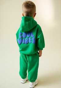 Gyerek zöld kapucnis pulóverben és melegítőnadrágban, amelynek hátulján "COOL VIBES" felirat látható, háttal áll világos háttér előtt.