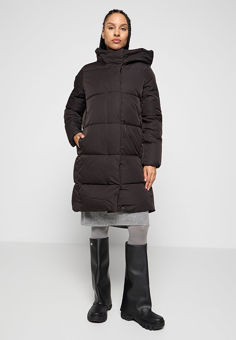 Frau in einem langen schwarzen gefütterten Mantel, grauem Rock, grauen Strumpfhosen und schwarzen kniehohen Stiefeln, die vor einem weißen Hintergrund steht.