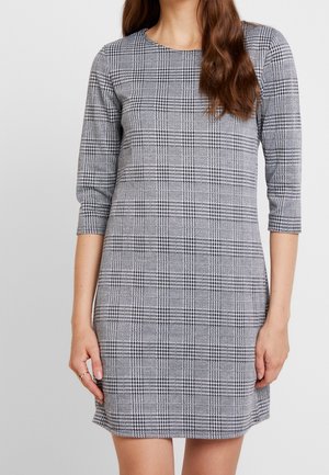 Femme portant une robe à carreaux grise et noire près du genou avec des manches trois-quarts, debout devant un fond clair uni.