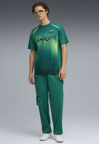 Grünes Sporthemd mit Farbverlauf und schwarzem "AMR25"-Druck, kombiniert mit passenden grünen Hosen mit Taschen und lockerer Passform, gekrönt von gelben Sonnenbrillen.