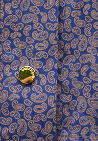 Granatowy materiał z powtarzającym się jasnobrązowym wzorem paisley. Posiada złoty guzik z tłoczonym tekstem, co wzbogaca szczegóły projektu.