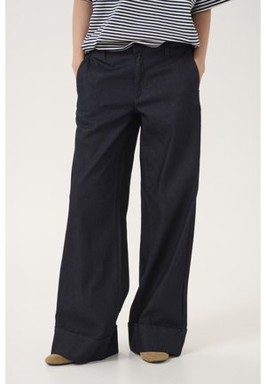 Personne portant un pantalon large foncé et une chemise rayée, les mains dans les poches, debout sur un fond clair.