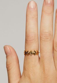 Anillo de oro con un patrón geométrico que presenta formas de diamante alternas y pequeños acentos negros; textura suave sobre un fondo de piel.