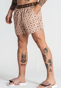 Costumi da bagno beige con un design a motivo che presenta loghi marroni. Vita elastica con una banda marrone, abbinati a ciabatte con dettagli logo.