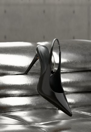 Glossy black high-heeled slingback shoe balanced on shiny silver metallic cushioned surface with smooth texture.