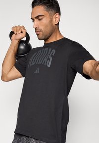 Svart kortermet T-skjorte med stor grå "ADIDAS"-logo, rund hals, lett stoff, kombinert med en kettlebell.