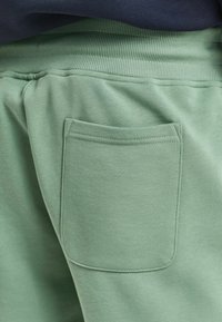 Pantalons de jogging verts avec une poche au dos, texture douce, taille côtelée et coupe détendue. Le tissu semble lisse et décontracté.