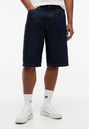 Mand iført mørkeblå denimshorts, hvide crew-sokker med logo og hvide sneakers, stående med den ene hånd i lommen.