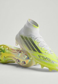 Ζευγάρι λευκών και κίτρινων ποδοσφαιρικών παπουτσιών με ψηλή κοπή, μαύρες ρίγες και σόλα με ολογραφικά καρφιά, εμφανιζόμενα σε ανοιχτό φόντο.