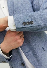 Jasnoniebieska lniana marynarka o teksturowanym wykończeniu. Posiada czarne guziki na mankietach, białą koszulę pod spodem oraz pojedynczą kieszeń na piersi.