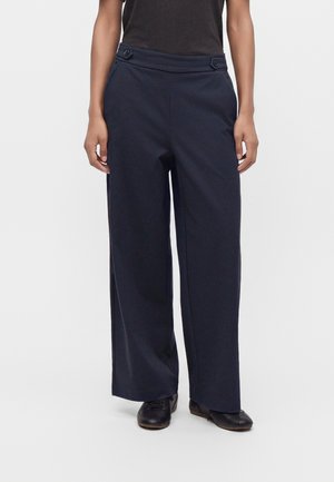 Person wearing high-waisted, wide-leg navy trousers with button details on the waistband and black shoes against a plain background.