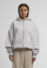 Femme portant un sweat zippé gris clair avec capuche et un pantalon beige ample, debout devant un fond gris uni.