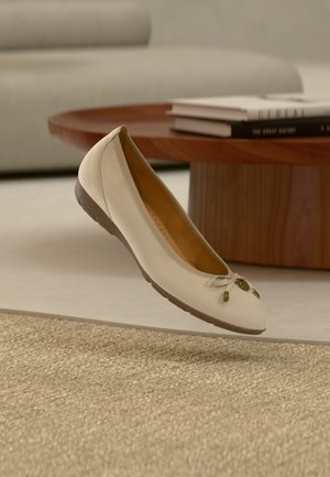 Beżowe płaskie buty z kokardą unoszące się nad teksturowanym dywanem w salonie z drewnianym stolikiem kawowym i ułożonymi książkami w tle.