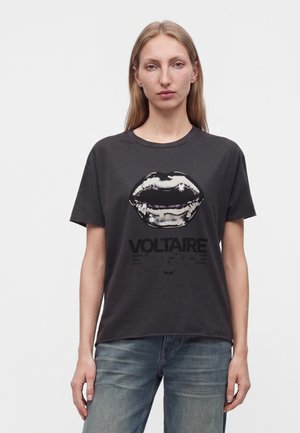 Jeune femme aux cheveux blonds raides portant un t-shirt noir avec un graphisme de lèvres brillantes et le mot "VOLTAIRE", associé à un jean bleu.