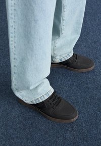 Czarne buty sportowe z perforowanym wzorem, z materiałowymi sznurówkami i gumową podeszwą, w połączeniu z jasnoniebieskimi dżinsami, które mają luźny krój.