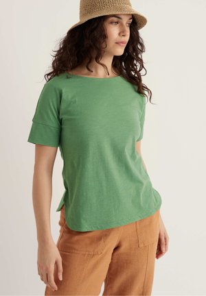 Kvinde med krøllet hår iført en beige flettet bøllehat, grøn kortærmet top og rustfarvede bukser, der ser til siden mod en lys baggrund.