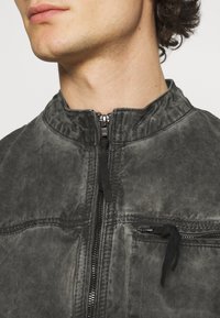 Närbild av en persons nedre ansikte och hals som bär en urblekt svart zip-up jacka med hög krage och dragkedja på bröstfickan.