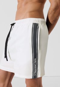 Fehér szövetből készült úszónadrág fekete oldalcsíkokkal. Gumis derékpánttal, zsinóros zárással és logóval ellátott részlettel rendelkezik az oldalon.