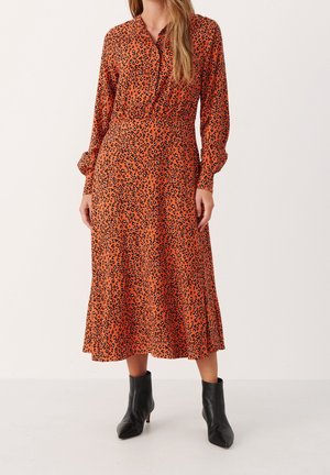 Kobieta w pomarańczowej sukience midi w czarny wzór w panterkę, z długimi rękawami, kołnierzykiem na guziki, w połączeniu z czarnymi botkami za kostkę.