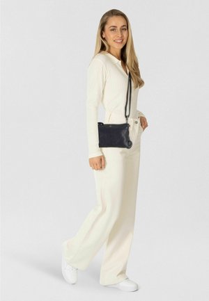 Jonge vrouw in een crème outfit, witte sneakers dragend en een zwarte schoudertas met een ankerhanger bij zich, glimlachend naar de camera.