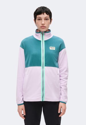Personne aux cheveux courts et foncés portant une veste polaire zippée bleu sarcelle et violet clair avec un petit logo sur la poitrine.